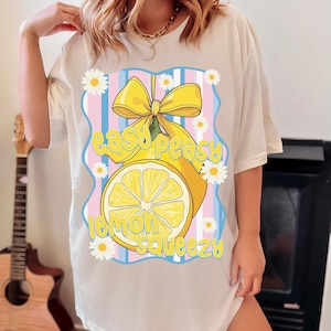 Pu&ograve; includere: Maglietta bianca con una grafica di un limone giallo con un fiocco giallo e il testo "easy peasy lemon squeezy" in lettere blu e rosa.