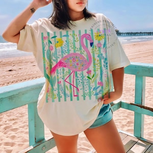 May include: Cream-colored t-shirt featuring a pink flamingo design with floral accents and a striped background. The flamingo is surrounded by colorful flowers and foliage. The shirt has short sleeves and a relaxed fit. Text on the shirt reads "TRAINING PROHIBITED ARWEN DIGITAL COPYRIGHTED".