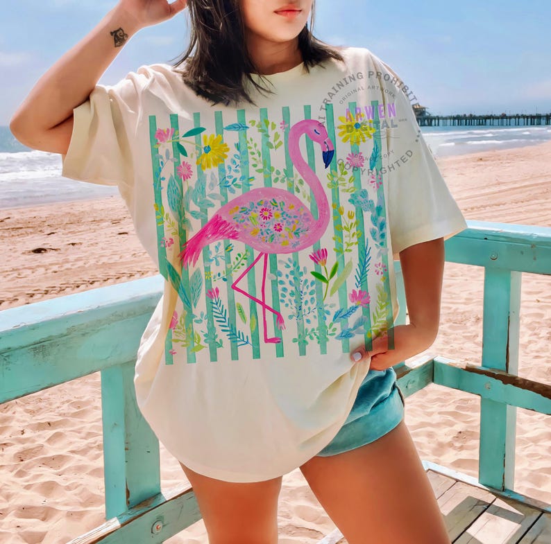 May include: Cream-colored t-shirt featuring a pink flamingo design with floral accents and a striped background. The flamingo is surrounded by colorful flowers and foliage. The shirt has short sleeves and a relaxed fit. Text on the shirt reads "TRAINING PROHIBITED ARWEN DIGITAL COPYRIGHTED".