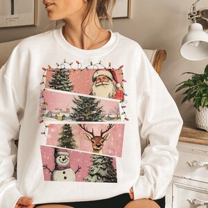 May include: White sweatshirt featuring a Christmas-themed graphic. The design includes Santa Claus, Christmas trees, a snowman, and a reindeer, all set against a pink backdrop. The graphic is framed with string lights, creating a festive look.