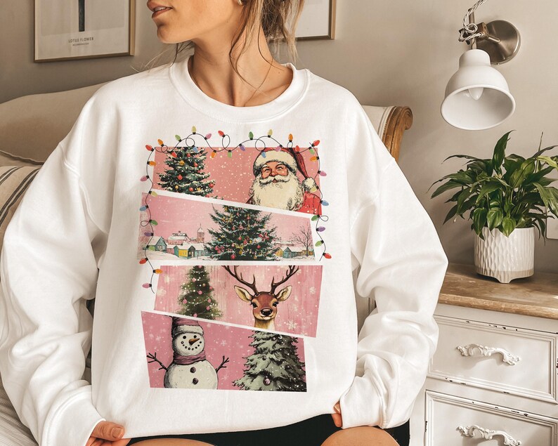 May include: White sweatshirt featuring a Christmas-themed graphic. The design includes Santa Claus, Christmas trees, a snowman, and a reindeer, all set against a pink backdrop. The graphic is framed with string lights, creating a festive look.