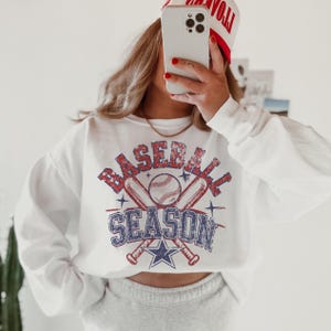 May include: White sweatshirt with a vintage-style graphic of crossed baseball bats and a baseball with the text "BASEBALL SEASON" in red, blue, and white.