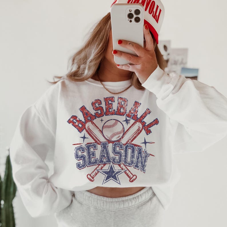 May include: White sweatshirt with a vintage-style graphic of crossed baseball bats and a baseball with the text "BASEBALL SEASON" in red, blue, and white.