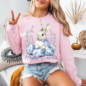 May include: Pink Easter-themed sweatshirt with a bunny and blue eggs design. The graphic features a white bunny with a blue bow, sitting on blue patterned eggs, with the words "Happy Easter" below. The sweatshirt is a light pink color.