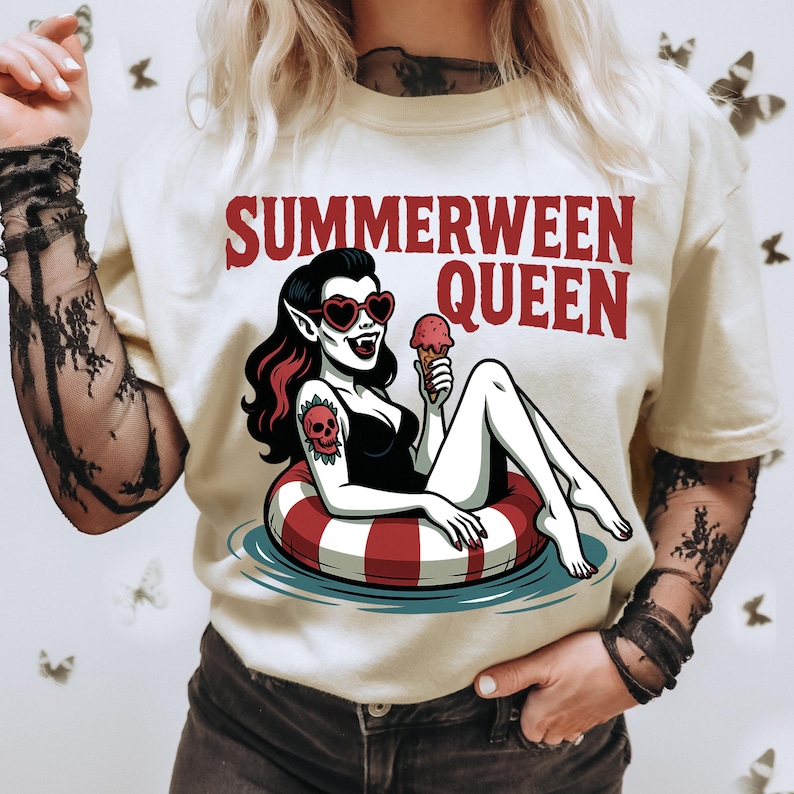 May include: Cream-colored t-shirt with the text "SUMMERWEEN QUEEN" in red. The graphic features a cartoon vampire with heart-shaped sunglasses, a skull tattoo, and an ice cream cone, lounging in a red and white striped inner tube.