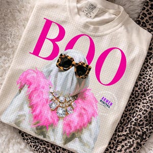 May include: A cream-colored t-shirt featuring a graphic of a ghost wearing leopard print sunglasses and a large, sparkling rhinestone statement necklace. The ghost is draped in a bright pink feather boa. The word BOO is printed in large, hot pink letters.