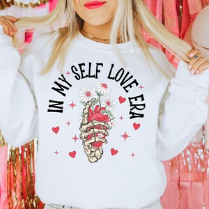 May include: White sweatshirt with the text "IN MY SELF LOVE ERA" in black. The graphic features a skeleton hand holding a heart with flowers. Red hearts and stars surround the graphic. A casual, comfortable top.