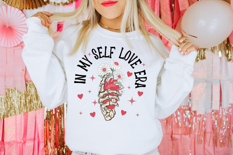 May include: White sweatshirt with the text "IN MY SELF LOVE ERA" in black. The graphic features a skeleton hand holding a heart with flowers. Red hearts and stars surround the graphic. A casual, comfortable top.
