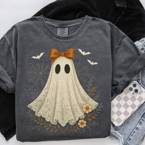 May include: Dark gray t-shirt featuring a ghost design with a brown bow, bats, and floral accents. The ghost has a lace-like appearance. The shirt is paired with blue jeans and a phone case with a checkered pattern. A black jacket is also visible.