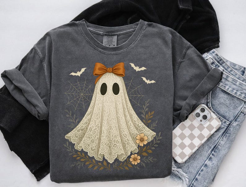 May include: Dark gray t-shirt featuring a ghost design with a brown bow, bats, and floral accents. The ghost has a lace-like appearance. The shirt is paired with blue jeans and a phone case with a checkered pattern. A black jacket is also visible.