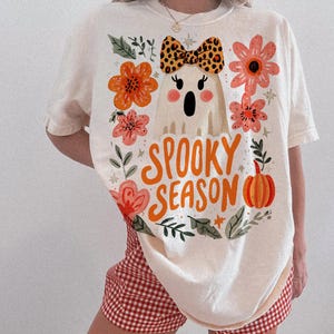 K&ouml;nnte beinhalten: Cremefarbenes T-Shirt mit einem Halloween-Motiv. Das Shirt zeigt einen Cartoon-Geist mit einer Leoparden-Schleife, umgeben von orangefarbenen und rosa Blumen, und die Worte "Spooky Season" in orangefarbenen Buchstaben. Das Shirt wird mit rot-wei&szlig; karierten Shorts kombiniert.