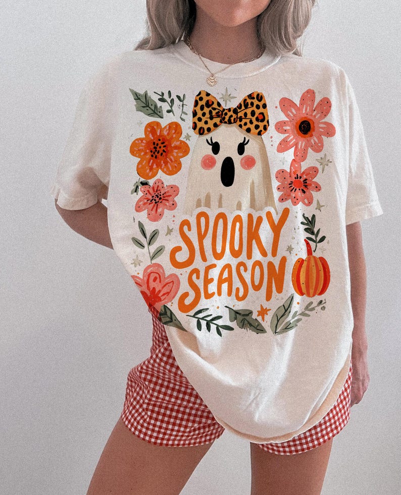K&ouml;nnte beinhalten: Cremefarbenes T-Shirt mit einem Halloween-Motiv. Das Shirt zeigt einen Cartoon-Geist mit einer Leoparden-Schleife, umgeben von orangefarbenen und rosa Blumen, und die Worte "Spooky Season" in orangefarbenen Buchstaben. Das Shirt wird mit rot-wei&szlig; karierten Shorts kombiniert.