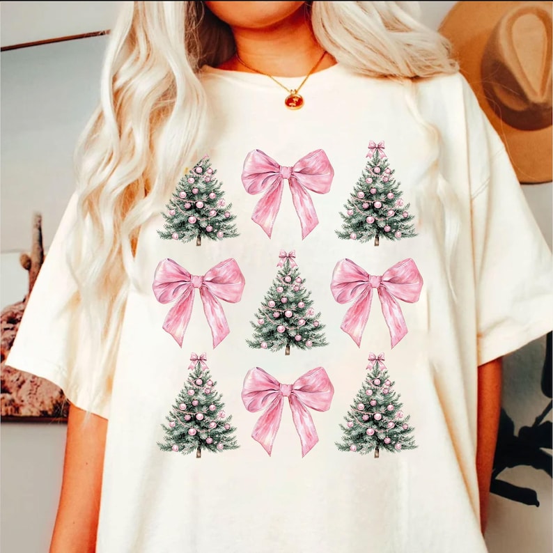 May include: Cream-coloured t-shirt with a pattern of pink bows and Christmas trees. The trees are green with pink ornaments. The bows are a solid pink colour. The shirt has a relaxed fit.