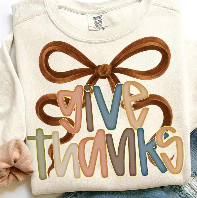May include: Cream-colored sweatshirt with the words "GIVE THANKS" in colorful, outlined letters. A large brown bow graphic is above the text. The sweatshirt is a Comfort Colors brand.