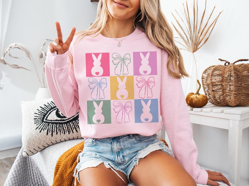 May include: A pink sweatshirt with a graphic design of pastel colored bunnies and bows in a grid pattern.
