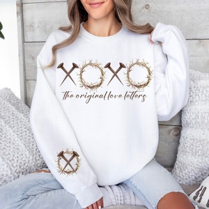 May include: White sweatshirt with a graphic of a crown of thorns and nails forming the letters "XOXO" with the text "The original love letters" below.