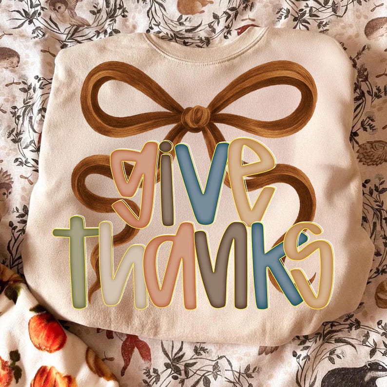 May include: Cream-colored sweatshirt with the words "GIVE THANKS" in colorful, outlined letters. A large brown bow graphic is above the text. The sweatshirt is displayed on a patterned fabric with fall-themed illustrations.
