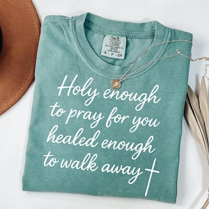 May include: A teal Comfort Colors t-shirt with the text "Holy enough to pray for you healed enough to walk away" in white script. A gold necklace with a pendant and a brown hat are also visible.