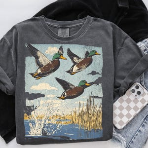 May include: A dark gray, crew neck t-shirt featuring a graphic of three ducks in flight over a body of water. The ducks have brown, white, and green feathers. The background includes blue sky, clouds, and reeds. A phone with a checkered case rests nearby.
