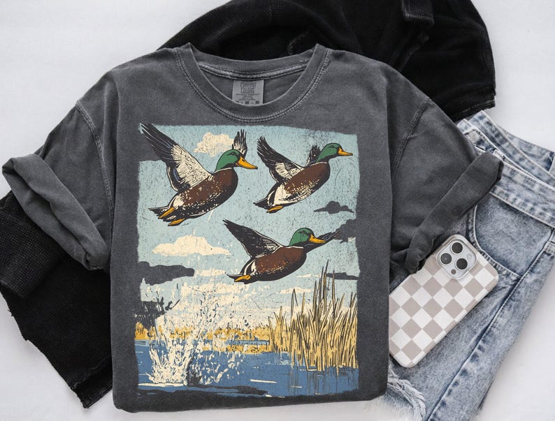 May include: A dark gray, crew neck t-shirt featuring a graphic of three ducks in flight over a body of water. The ducks have brown, white, and green feathers. The background includes blue sky, clouds, and reeds. A phone with a checkered case rests nearby.