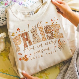 May include: A cream-coloured sweatshirt featuring the text "I will PRAISE Him in every season" and "PSALM 146:2". The word "PRAISE" is designed with floral and plaid patterns. Autumn leaves and floral accents are printed around the text.