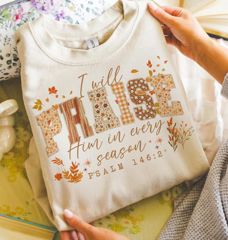 May include: A cream-coloured sweatshirt featuring the text "I will PRAISE Him in every season" and "PSALM 146:2". The word "PRAISE" is designed with floral and plaid patterns. Autumn leaves and floral accents are printed around the text.