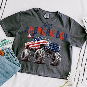 May include: Dark gray t-shirt with the text "All American Boy" and a graphic of a monster truck decorated with the American flag. The shirt is paired with blue jeans and a green and white hair clip.