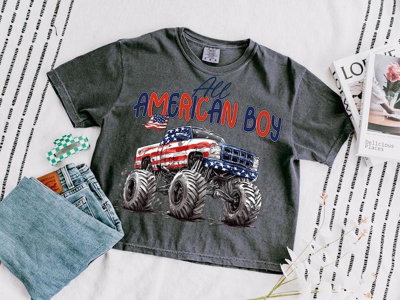 May include: Dark gray t-shirt with the text "All American Boy" and a graphic of a monster truck decorated with the American flag. The shirt is paired with blue jeans and a green and white hair clip.