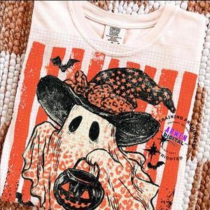May include: A cream-coloured t-shirt featuring a graphic of a ghost wearing a witch hat with a leopard print pattern. The ghost holds a black jack-o'-lantern bucket. The background design includes orange vertical stripes and a grid pattern.