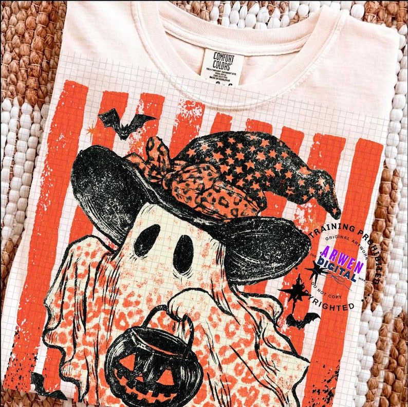 May include: A cream-coloured t-shirt featuring a graphic of a ghost wearing a witch hat with a leopard print pattern. The ghost holds a black jack-o'-lantern bucket. The background design includes orange vertical stripes and a grid pattern.