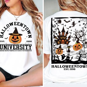 May include: White t-shirt with a Halloween design. The front displays a pumpkin in a witch's hat, the words "Halloweentown University", and the year 1998. The back shows a haunted castle, pumpkins, bats, and the text "Halloweentown Est. 1998".