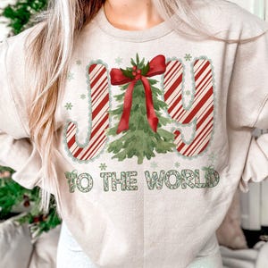Puede incluir: Sudadera beige con un gr&aacute;fico navide&ntilde;o. El dise&ntilde;o presenta las palabras "JOY TO THE WORLD" en rayas rojas y blancas de bast&oacute;n de caramelo y letras verdes, con un &aacute;rbol de Navidad y copos de nieve.