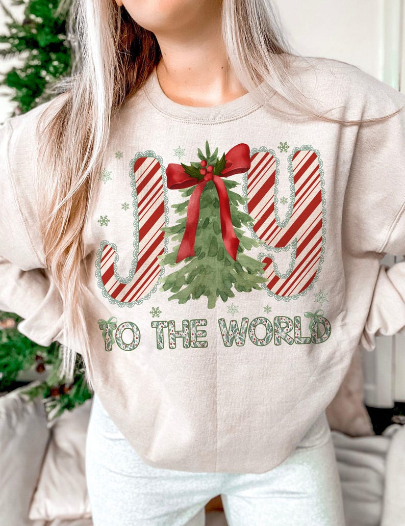 Puede incluir: Sudadera beige con un gr&aacute;fico navide&ntilde;o. El dise&ntilde;o presenta las palabras "JOY TO THE WORLD" en rayas rojas y blancas de bast&oacute;n de caramelo y letras verdes, con un &aacute;rbol de Navidad y copos de nieve.
