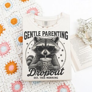 Puede incluir: Camiseta color crema con un mapache con gafas de sol y una taza. El texto dice "GENTLE PARENTING Dropout EST. THIS MORNING". La camiseta est&aacute; sobre una manta de ganchillo con motivos florales.