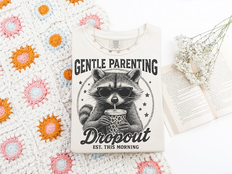 Puede incluir: Camiseta color crema con un mapache con gafas de sol y una taza. El texto dice "GENTLE PARENTING Dropout EST. THIS MORNING". La camiseta est&aacute; sobre una manta de ganchillo con motivos florales.