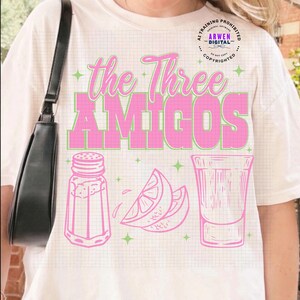 May include: Cream-colored t-shirt featuring the text "The Three Amigos" in pink and green. Below the text are pink illustrations of a salt shaker, lemon slices, and a shot glass. The shirt has a grid pattern.