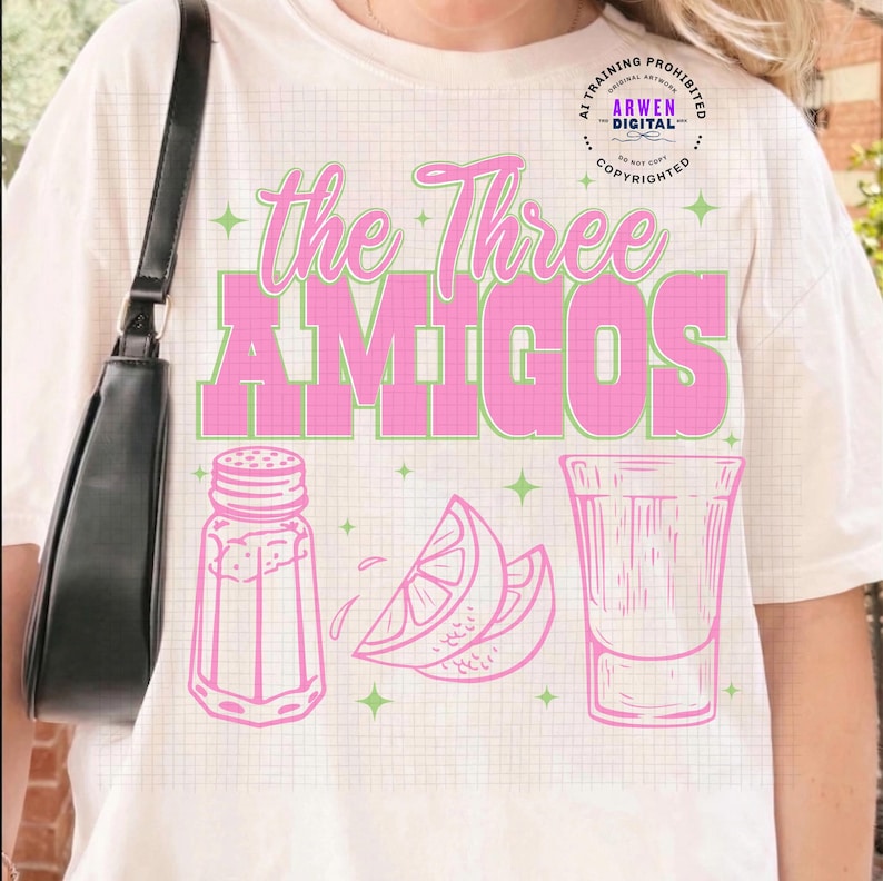 May include: Cream-colored t-shirt featuring the text "The Three Amigos" in pink and green. Below the text are pink illustrations of a salt shaker, lemon slices, and a shot glass. The shirt has a grid pattern.