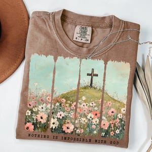 May include: A brown t-shirt with a painting of a field of flowers and a cross on a hill. The text "NOTHING IS IMPOSSIBLE WITH GOD" is printed on the bottom of the shirt.