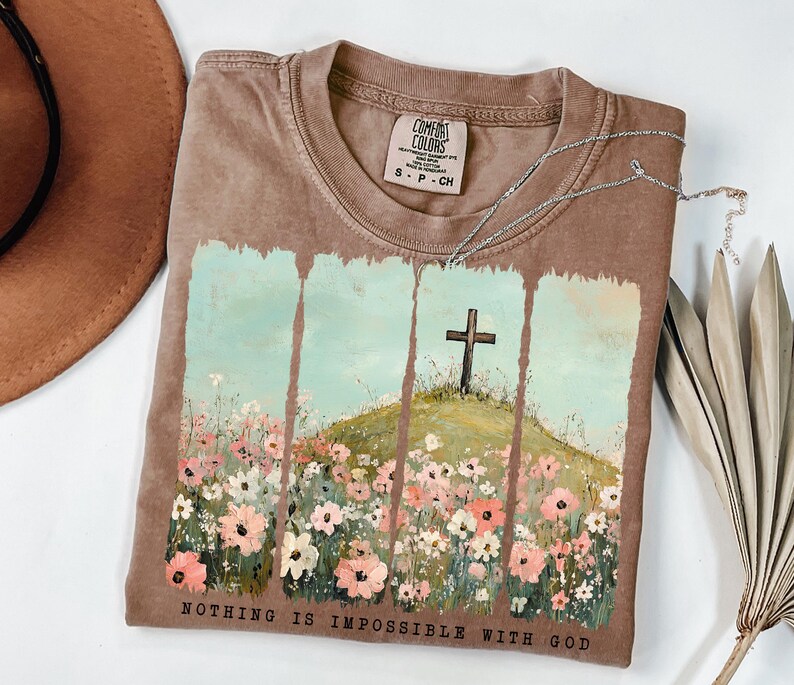 May include: A brown t-shirt with a painting of a field of flowers and a cross on a hill. The text "NOTHING IS IMPOSSIBLE WITH GOD" is printed on the bottom of the shirt.