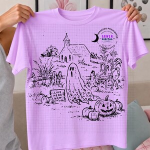 May include: A light purple t-shirt featuring a black line art graphic of a haunted scene. The design includes a ghost, a church, pumpkins, corn stalks, and a sign that reads "LIVE HERE NOW." A circular logo with the text "ARWEN DIGITAL" is visible.