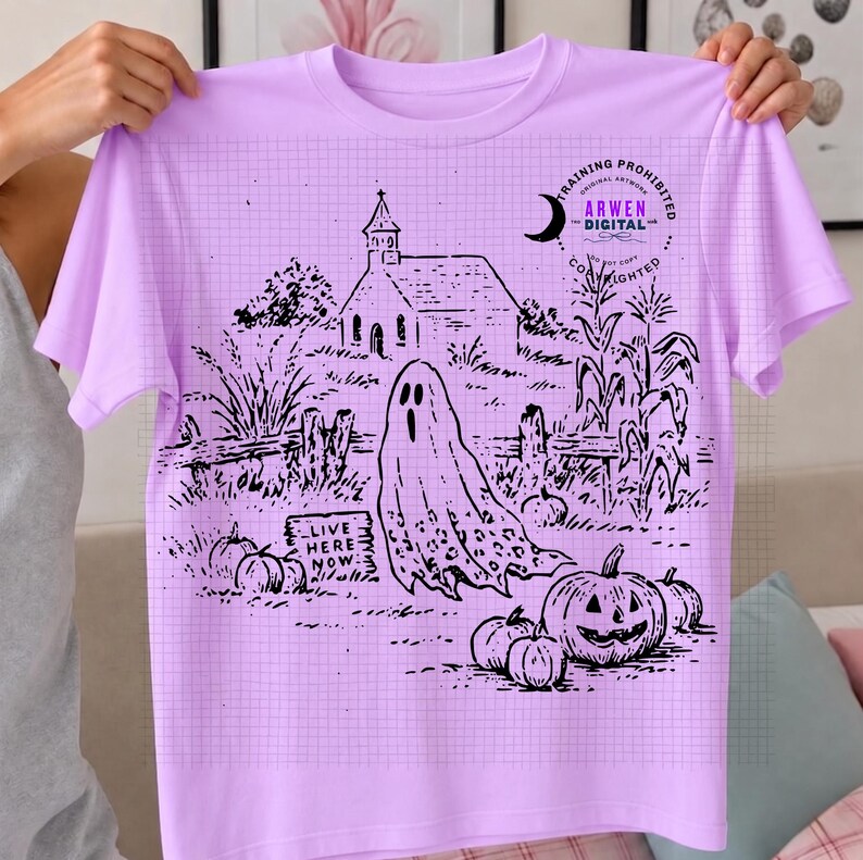 May include: A light purple t-shirt featuring a black line art graphic of a haunted scene. The design includes a ghost, a church, pumpkins, corn stalks, and a sign that reads "LIVE HERE NOW." A circular logo with the text "ARWEN DIGITAL" is visible.
