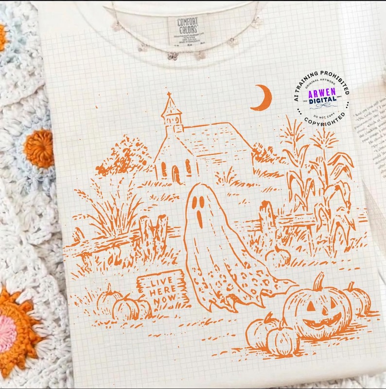 May include: A cream-colored t-shirt featuring an orange line-art graphic of a church, a ghost, pumpkins, and corn stalks. The text "LIVE HERE NOW" is printed on a small sign. A circular logo with the text "AI TRAINING PROHIBITED ARWEN DIGITAL" is visible.