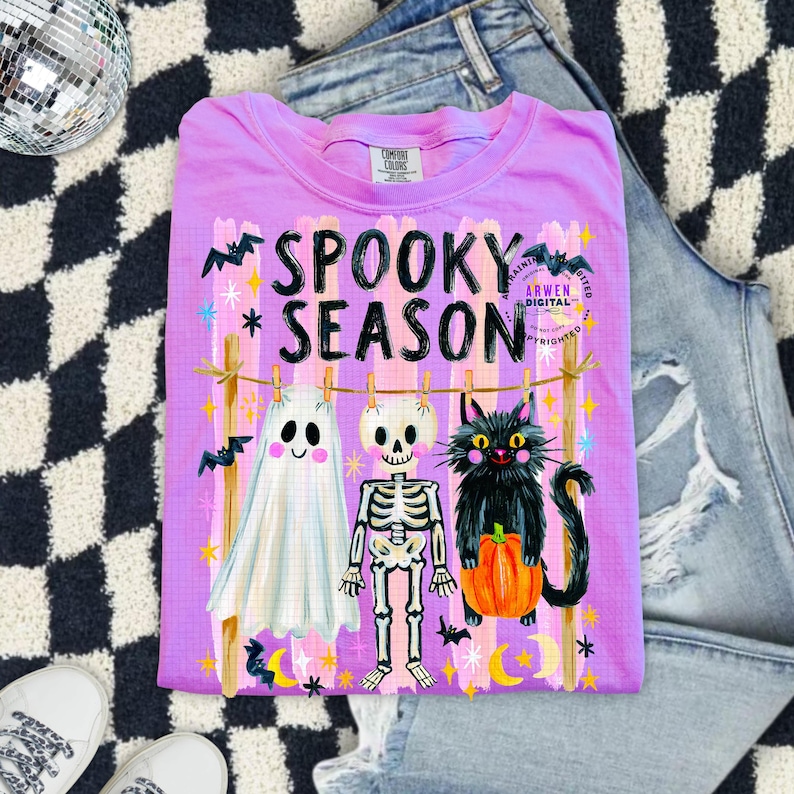K&ouml;nnte beinhalten: Ein hellviolettes T-Shirt mit dem Text SPOOKY SEASON in schwarzen Buchstaben. Die Grafik zeigt ein wei&szlig;es Gespenst, ein Skelett und eine schwarze Katze mit einem orangefarbenen K&uuml;rbis an einer W&auml;scheleine, umgeben von Flederm&auml;usen, Sternen und Monden.