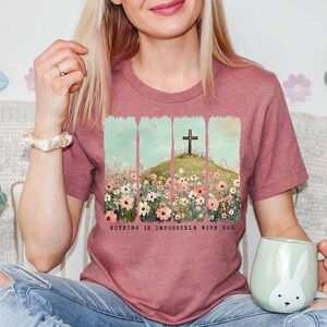 May include: A pink heather t-shirt with a floral design and a cross on a hill. The text "Nothing is impossible with God" is printed on the bottom of the shirt.