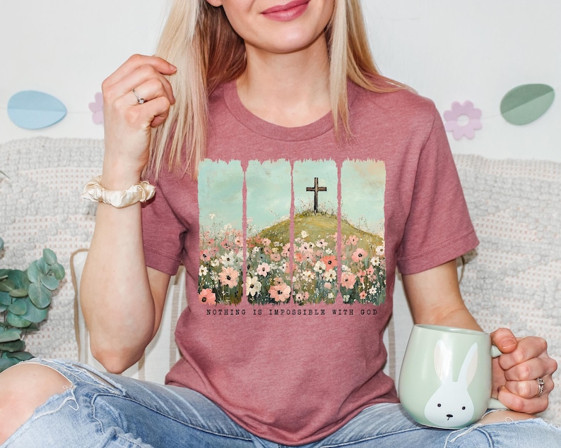 May include: A pink heather t-shirt with a floral design and a cross on a hill. The text "Nothing is impossible with God" is printed on the bottom of the shirt.