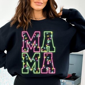May include: Black sweatshirt with the word "MAMA" in large, neon-style letters. The letters are outlined in green and pink, with small, colorful lights filling the inside of each letter. The sweatshirt is a casual, comfortable style.