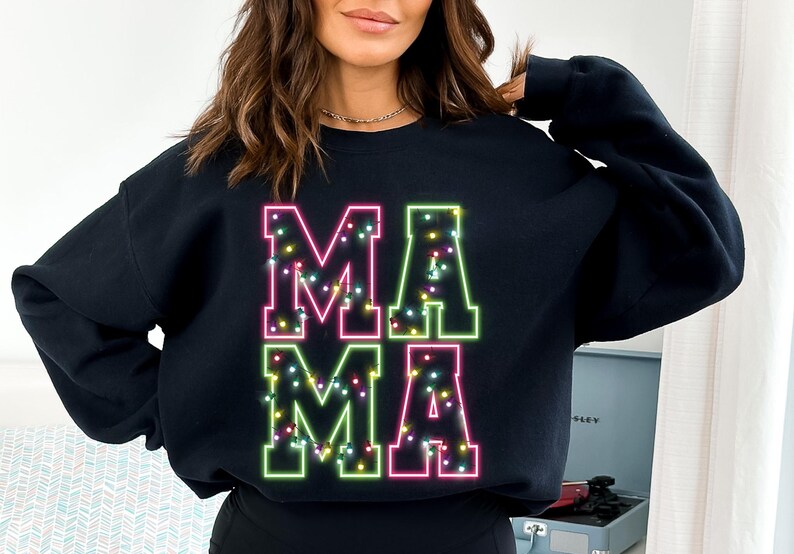 May include: Black sweatshirt with the word "MAMA" in large, neon-style letters. The letters are outlined in green and pink, with small, colorful lights filling the inside of each letter. The sweatshirt is a casual, comfortable style.