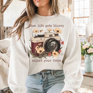 May include: White sweatshirt with a vintage camera graphic and the text "When life gets blurry adjust your focus". The camera is surrounded by flowers in shades of yellow, red, and brown.