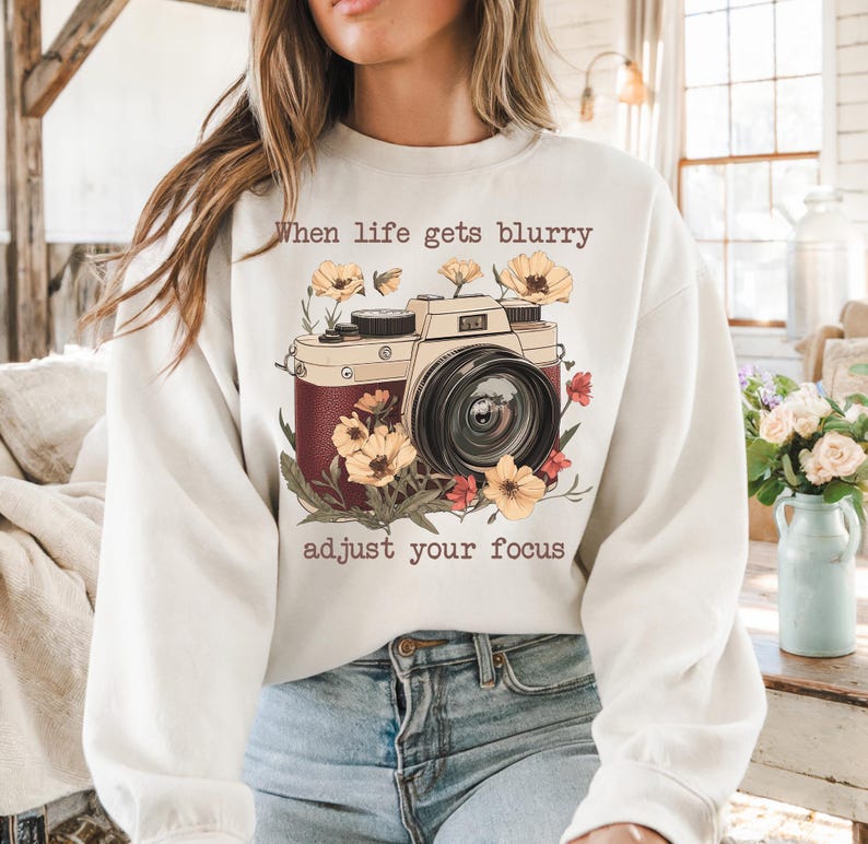 May include: White sweatshirt with a vintage camera graphic and the text "When life gets blurry adjust your focus". The camera is surrounded by flowers in shades of yellow, red, and brown.