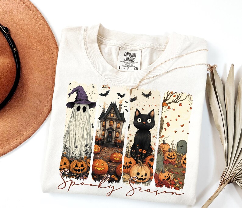 May include: Cream-colored t-shirt featuring a Halloween-themed design. The design includes a ghost wearing a witch hat, a haunted house, and a black cat surrounded by pumpkins. The words "Spooky Season" are written in a script font.